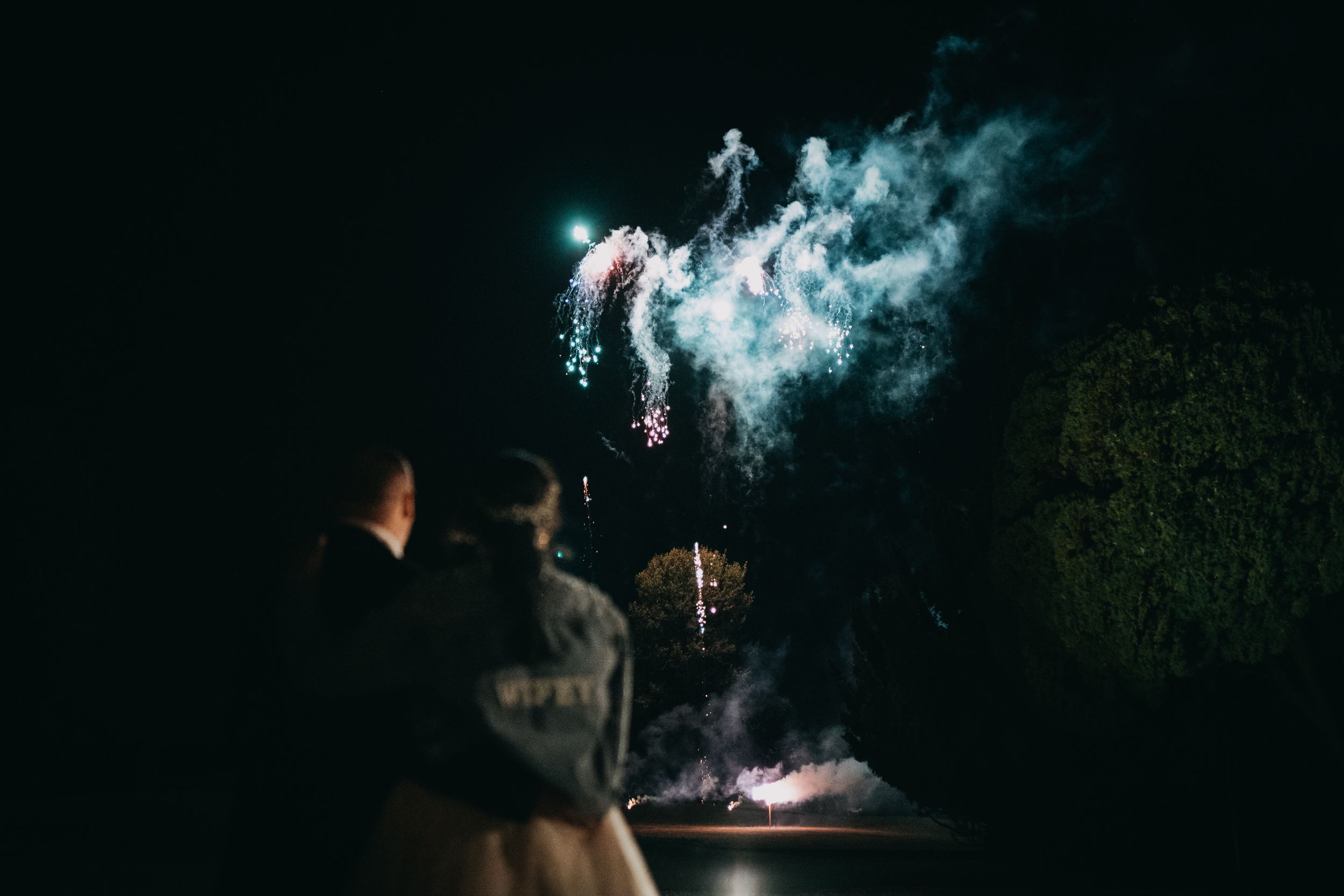 Wedding couple watching colorful fireworks exploding in the night sky at Holmewood Hall with bright turquoise bursts against dark foliage.