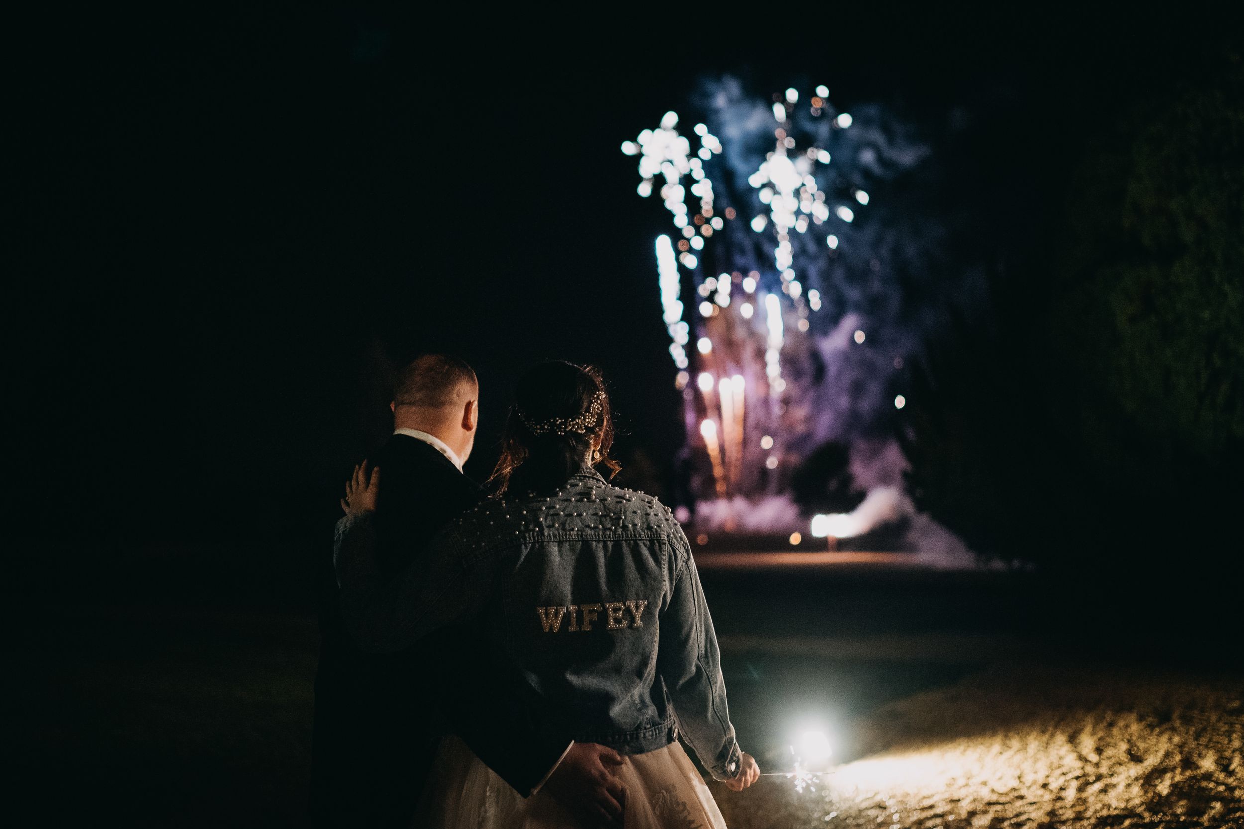 Two people watch colorful fireworks light up the night sky from a dark viewing area.