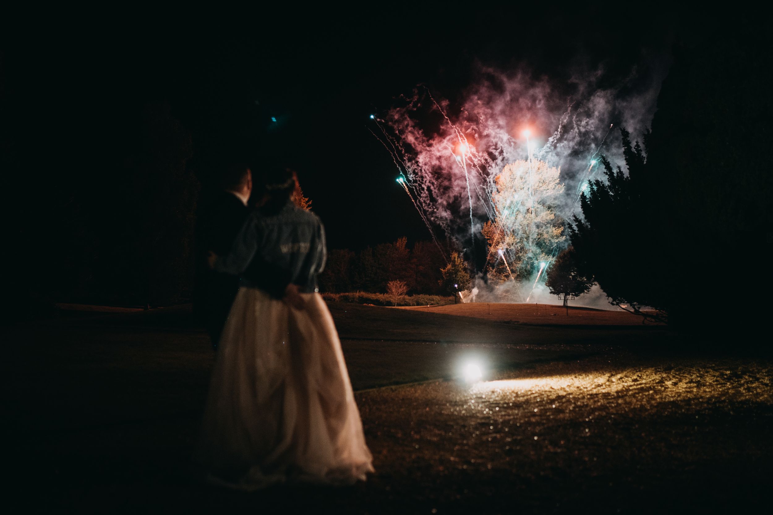 A couple silhouetted against bright fireworks display on a beach at night during a wedding celebration.