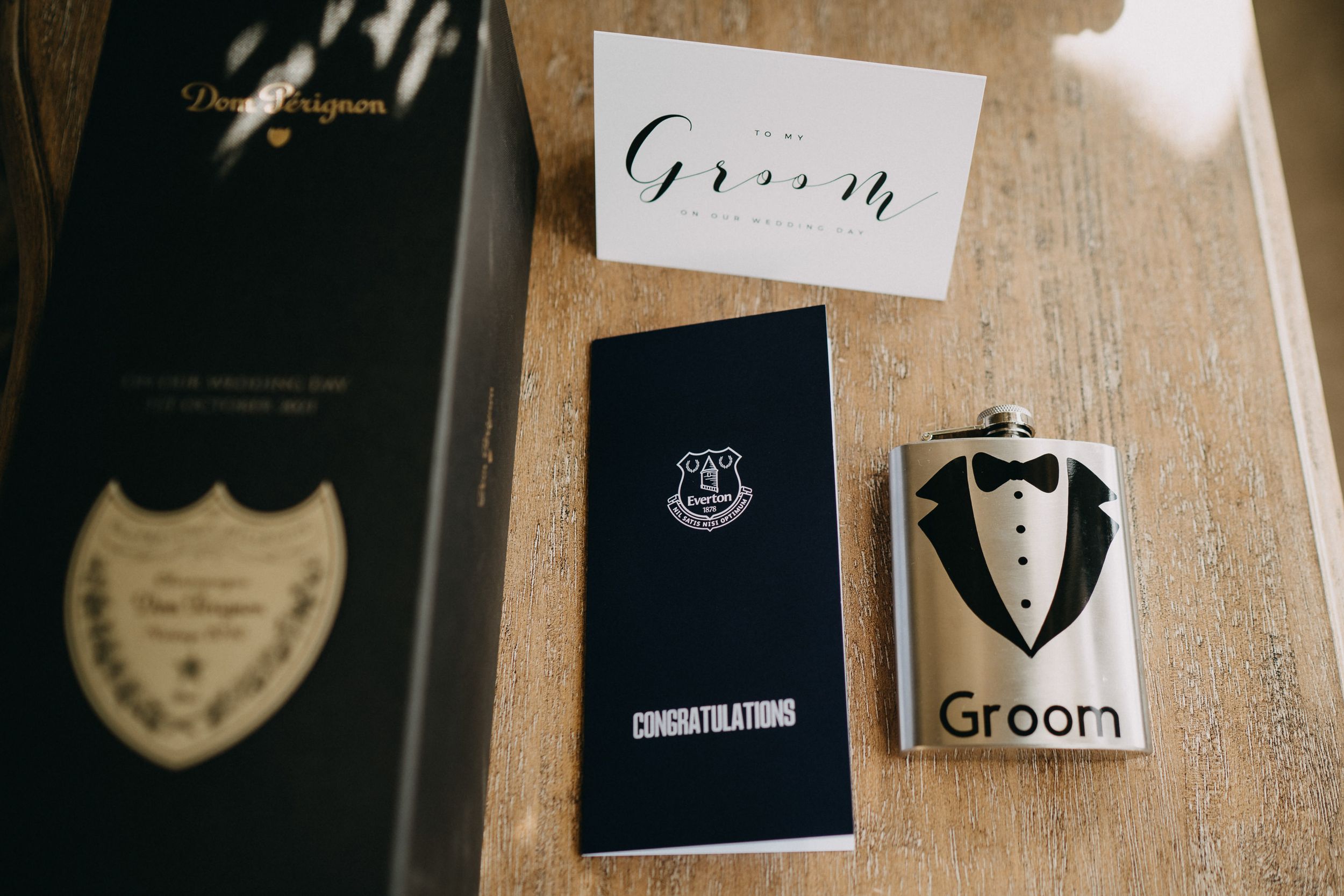 Wedding accessories including a groom flask, congratulations card, and Dom Perignon champagne bottle on wooden surface.