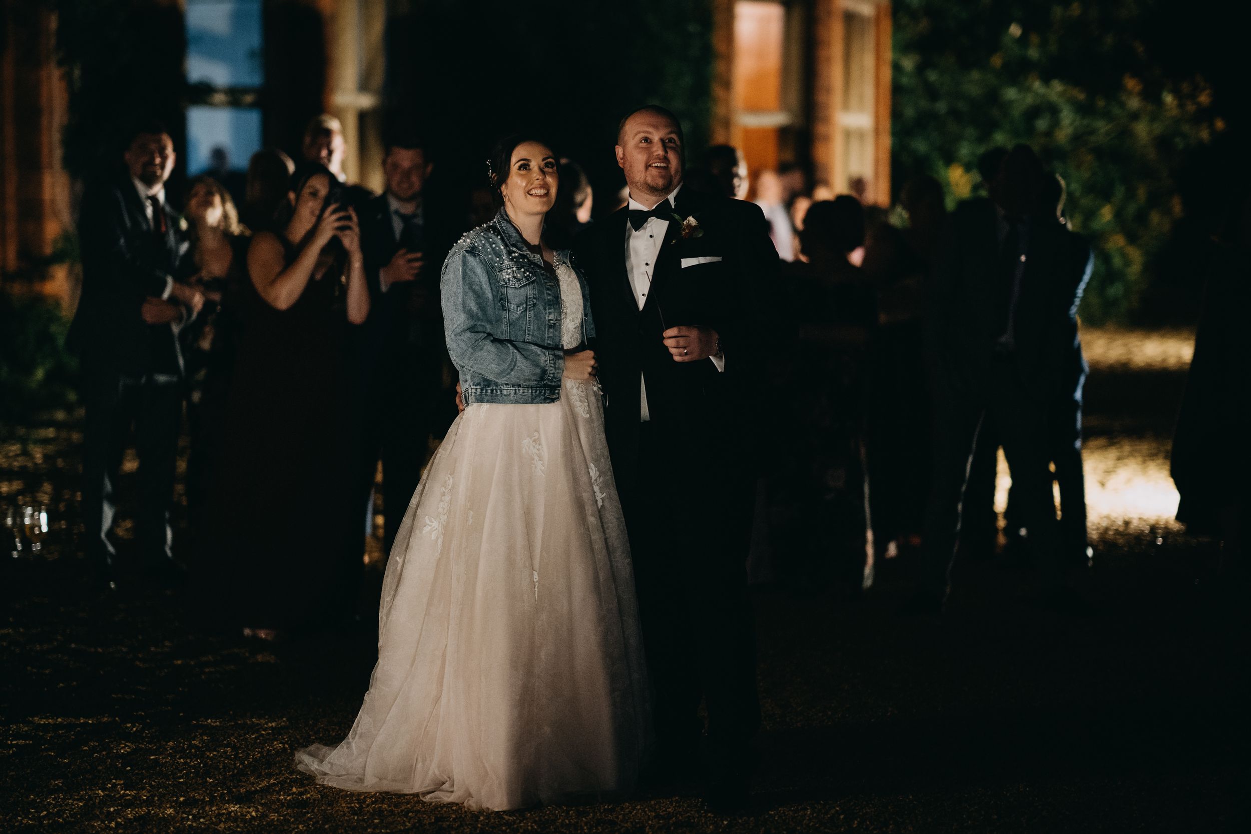 A wedding celebration at night with a bride wearing a denim jacket over her tulle gown alongside guests in formal attire.