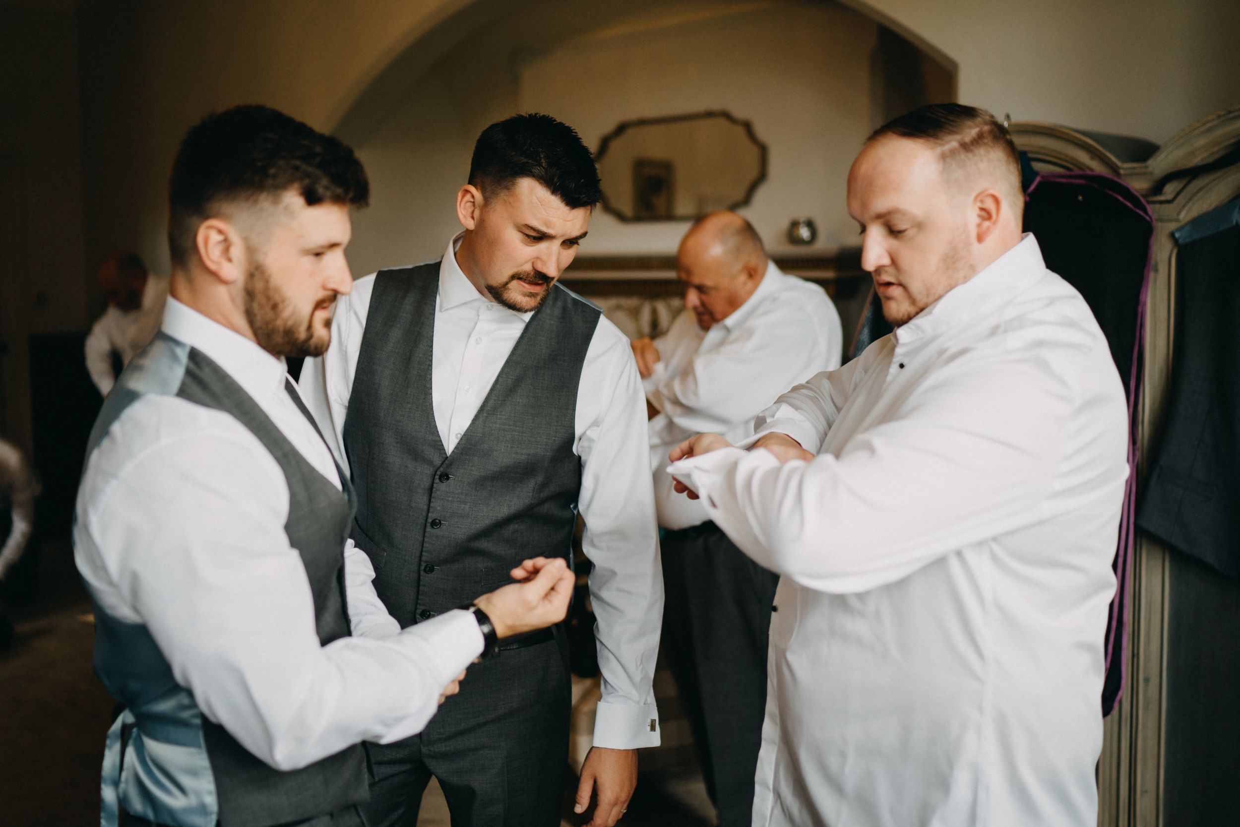 Groomsmen help each other get dressed and adjust their formal attire while preparing for a wedding ceremony.