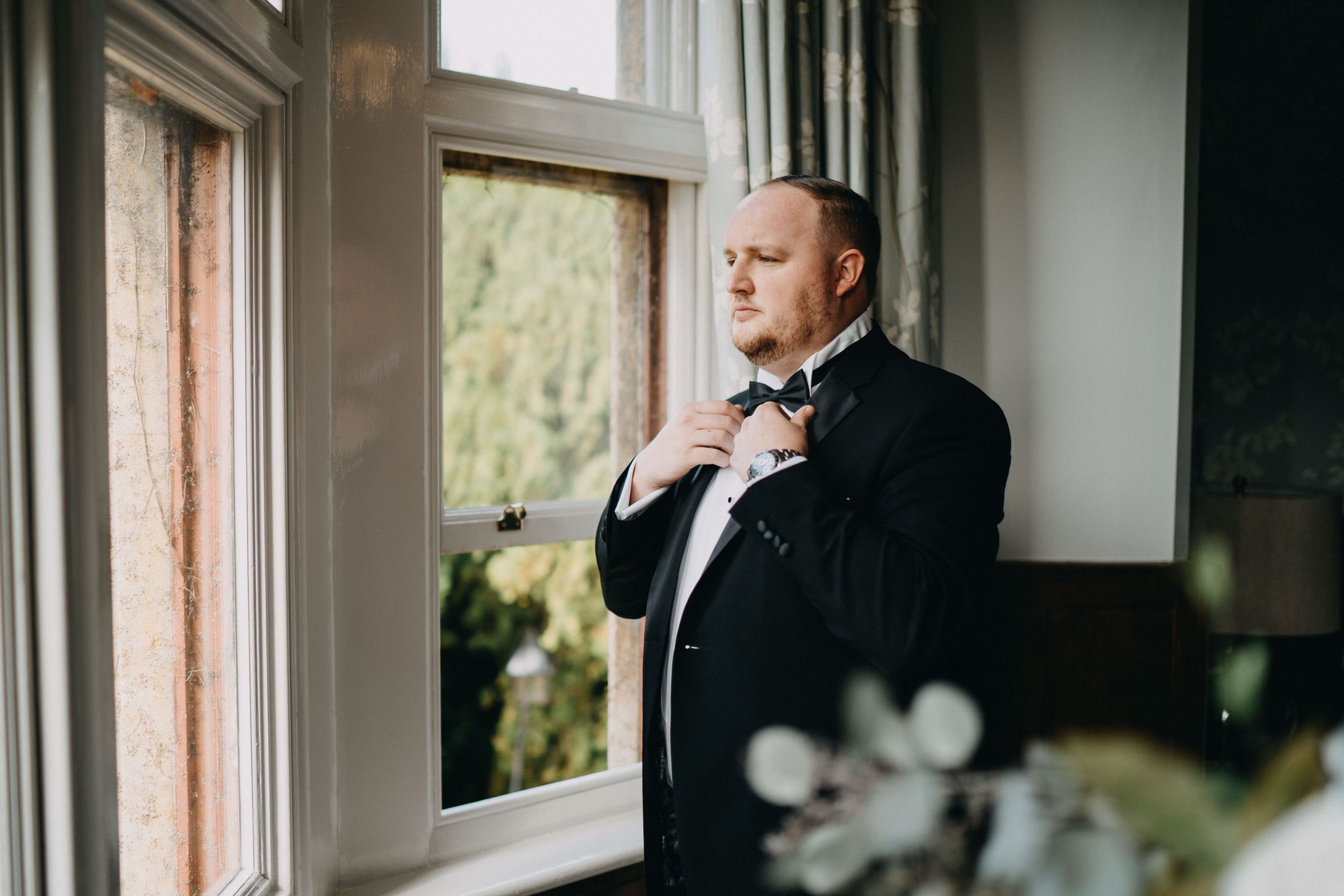 A person in a formal black tuxedo and bow tie adjusts their collar while standing by a window in natural light.