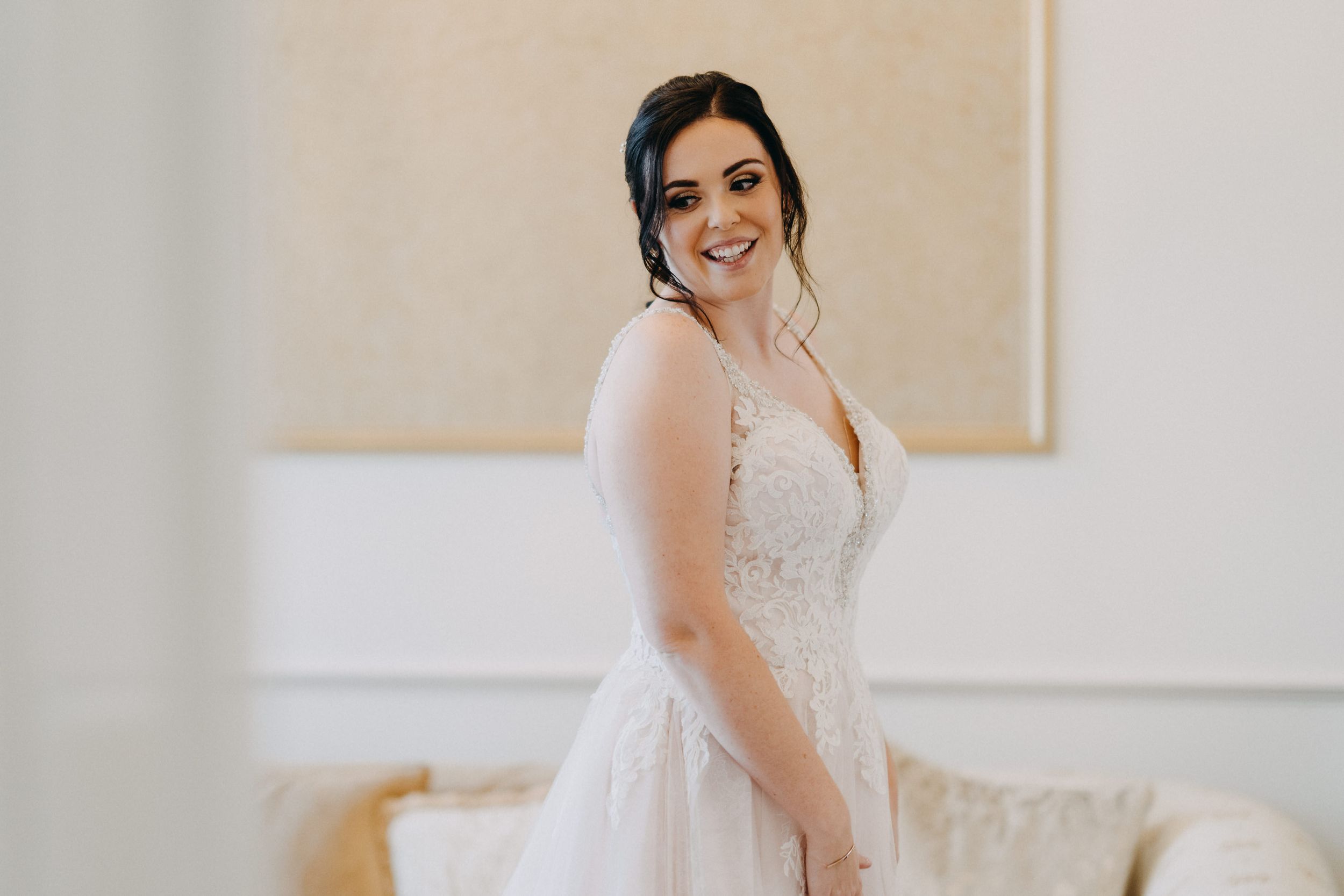 Bride wearing an elegant white lace wedding dress smiles warmly in a light-filled room.