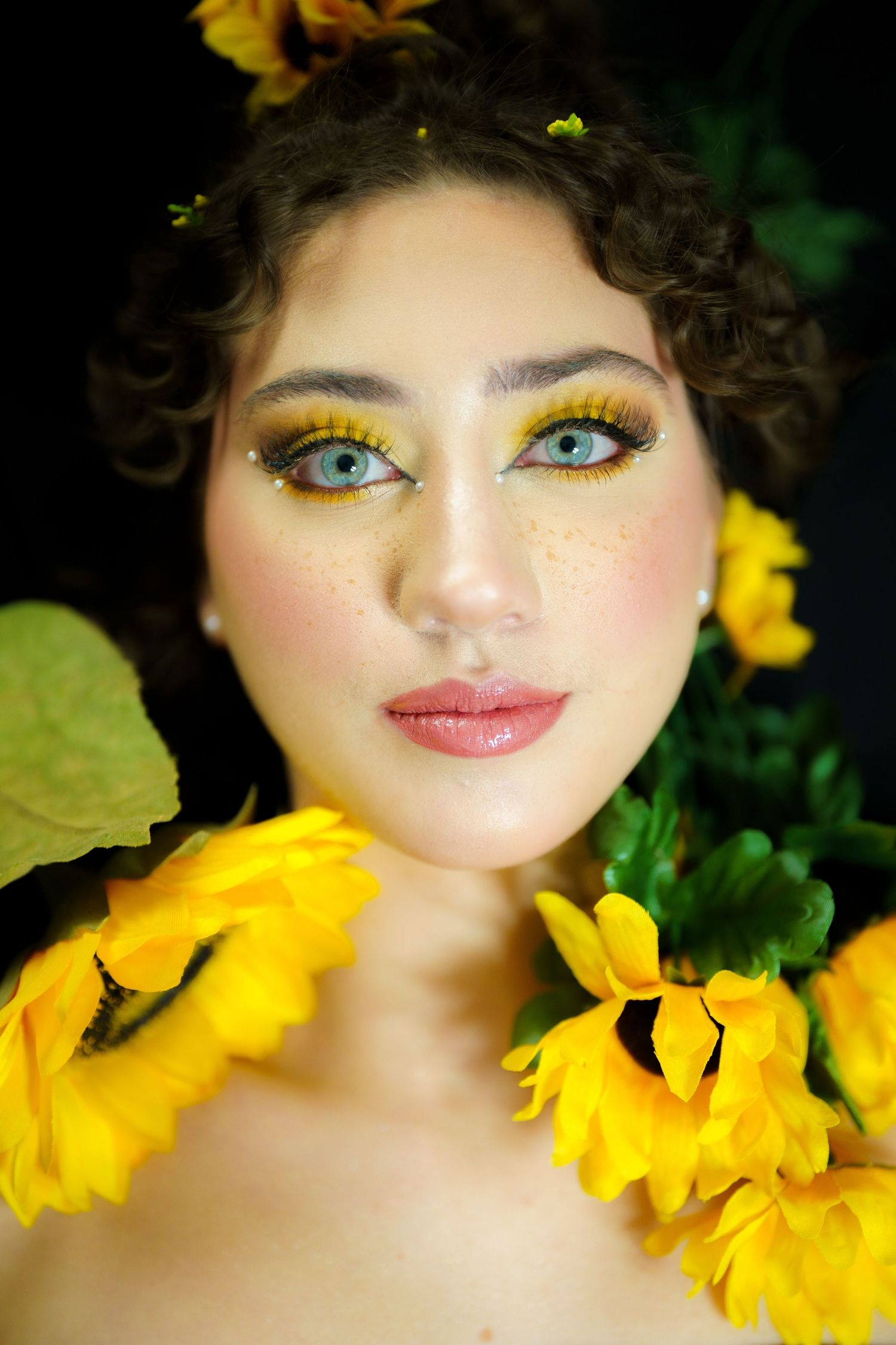 Portrait with vibrant yellow sunflowers showcasing glittery gold eyeshadow makeup and curly dark hair against greenery.