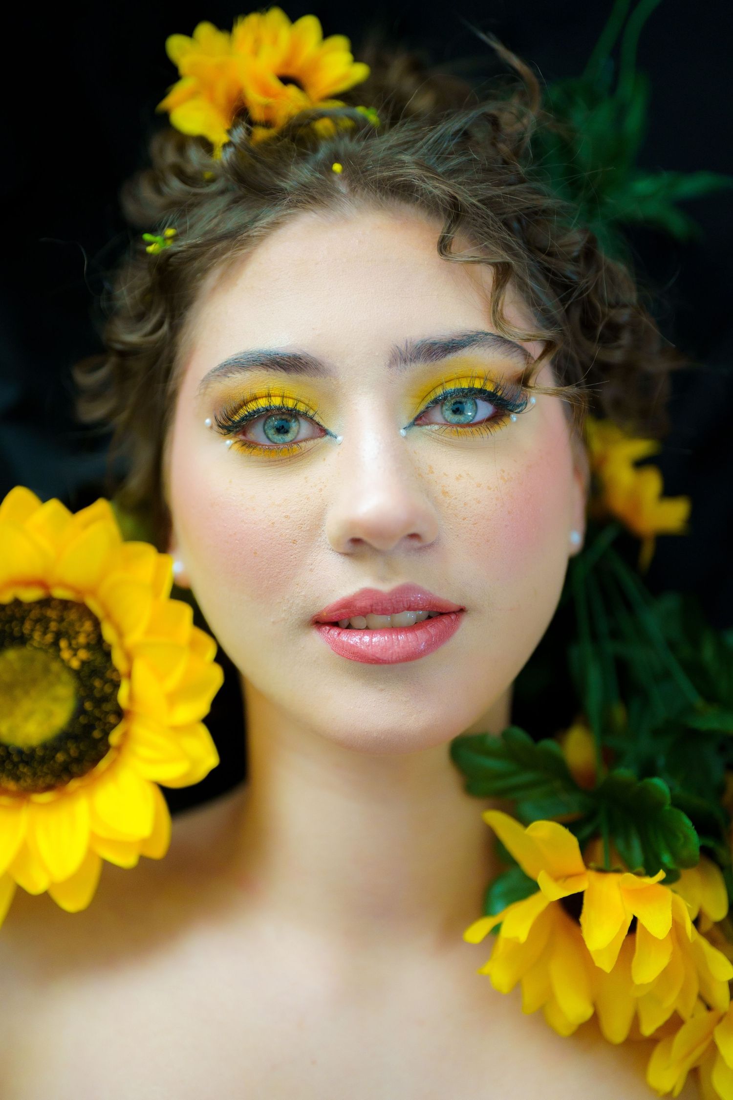 Artistic beauty portrait with vibrant yellow sunflowers and matching yellow eyeshadow makeup against dark background.