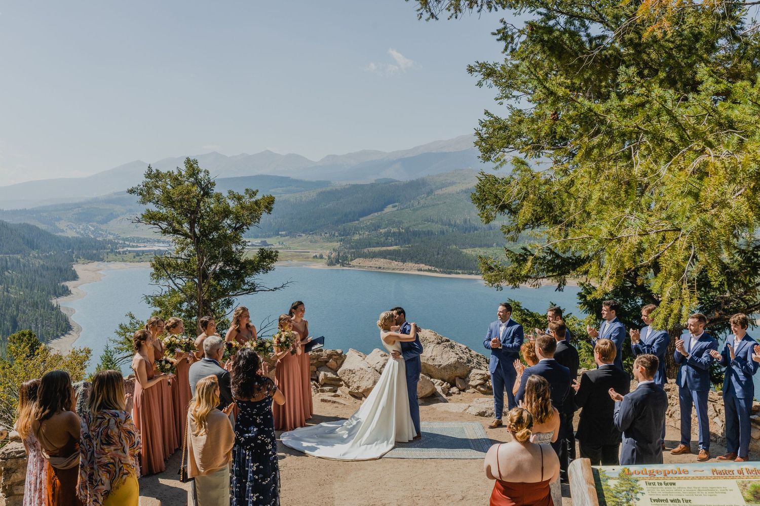 A scenic mountain wedding ceremony overlooking a pristine lake and rolling hills on a bright summer day.