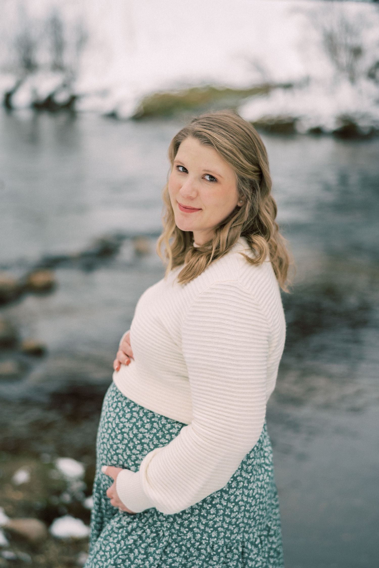 Winter maternity portrait by frozen river with woman in cream sweater and floral skirt.