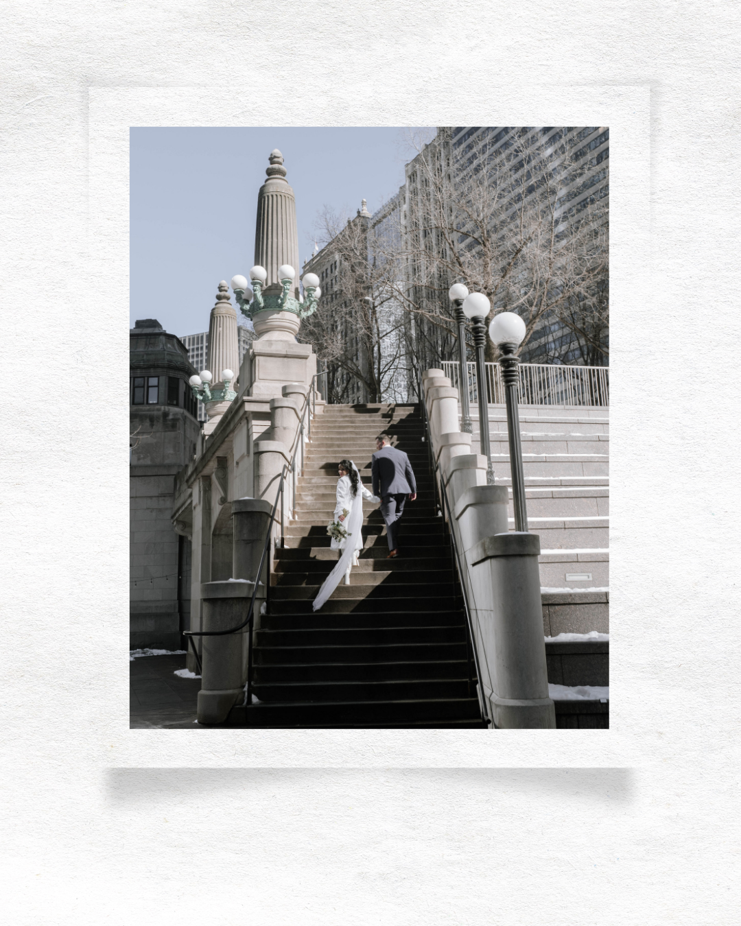 Two people ascend ornate white stone steps with decorative lampposts and columns leading up to a building on an urban campus.