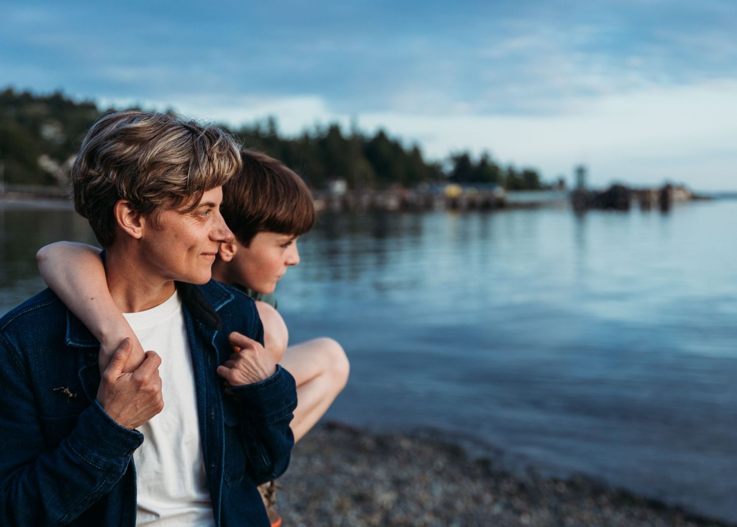 Figure sits with younger companion at waterfront during sunset, overlooking tranquil bay with forested shoreline in background.