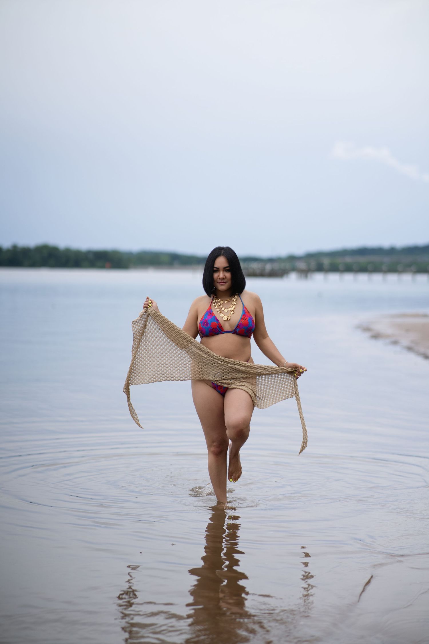 Person wearing purple bikini holding sheer fabric while standing in shallow calm water at beach during overcast day.