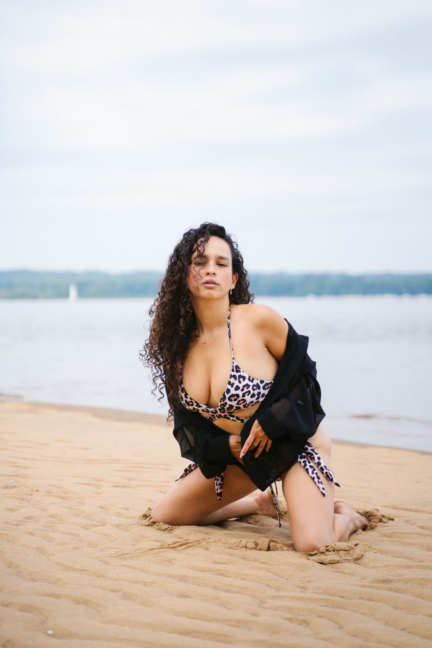 A person in a leopard print bikini and black jacket poses on a sandy beach with water and sailboats in the background.