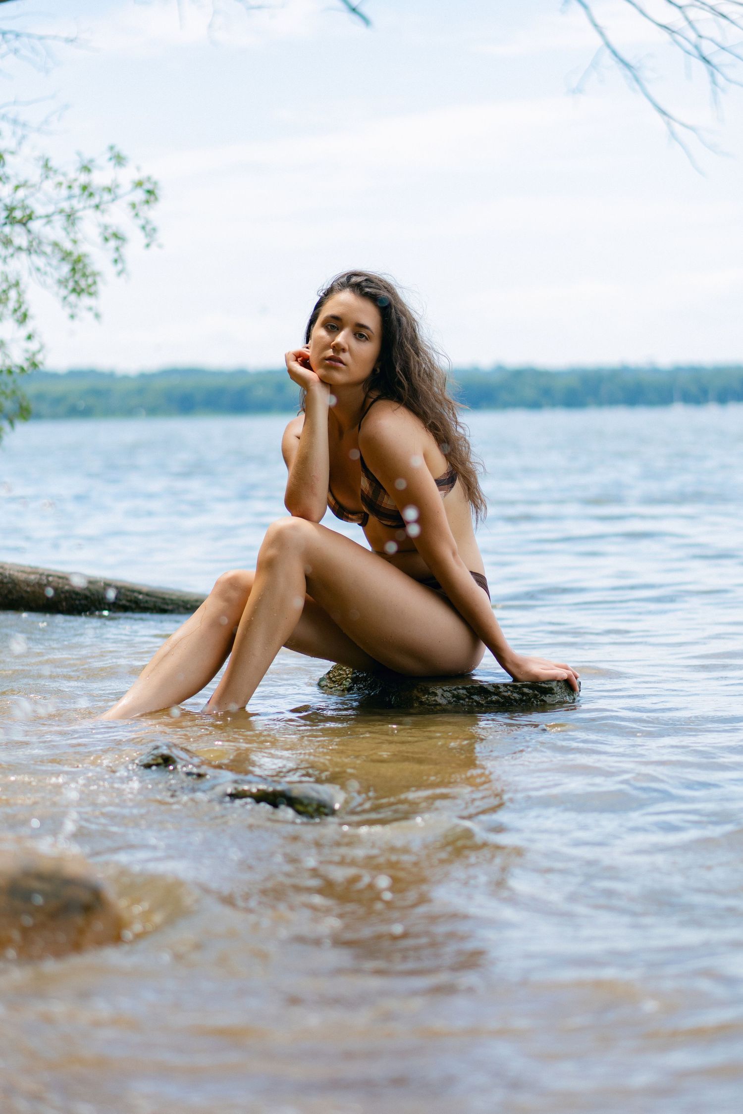 Person in a brown swimsuit poses thoughtfully on a rock at the edge of a serene lake on a summer day.