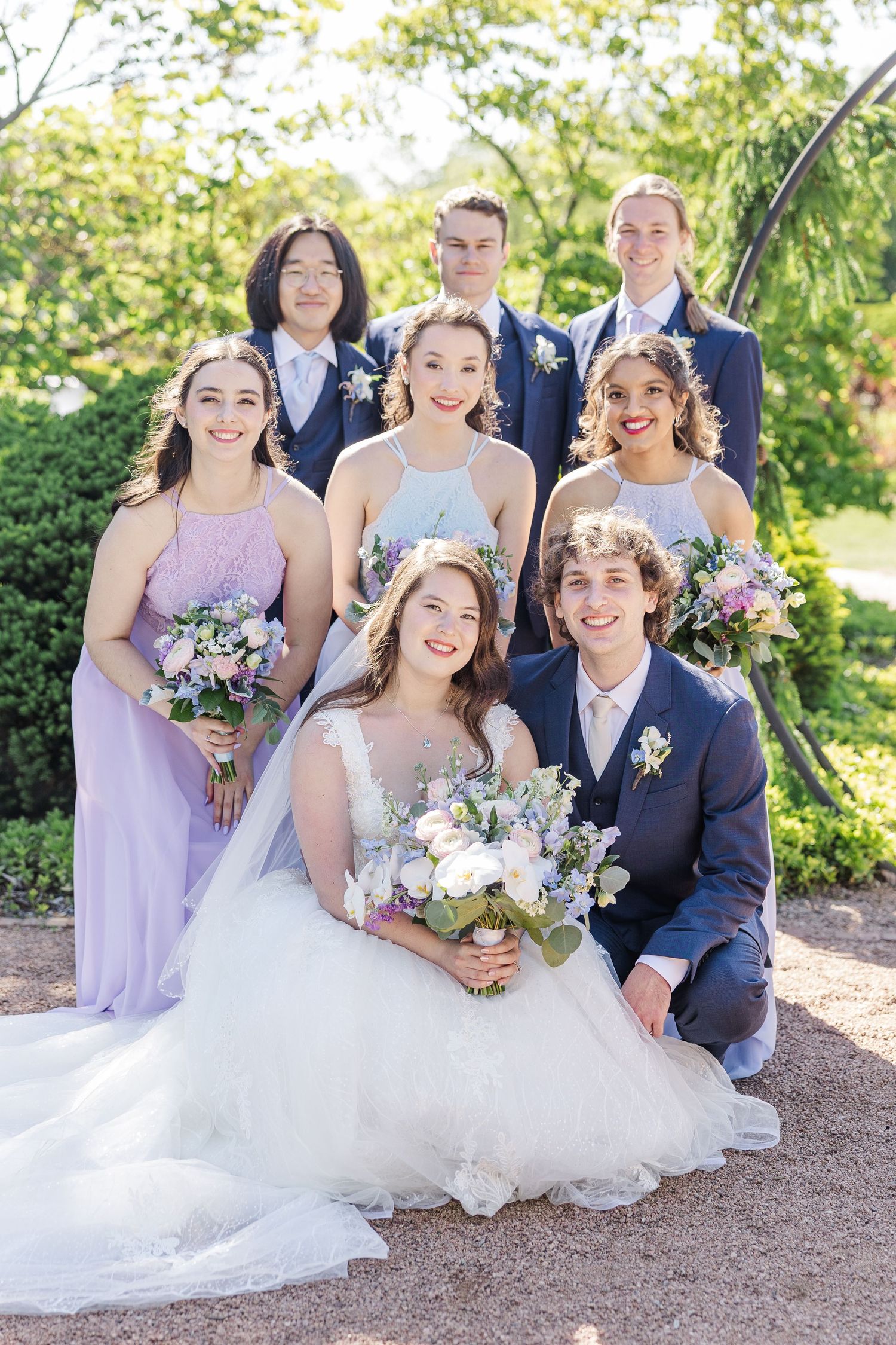 Wedding party poses together outdoors in pastel lilac attire with lush floral bouquets on a sunny spring day.