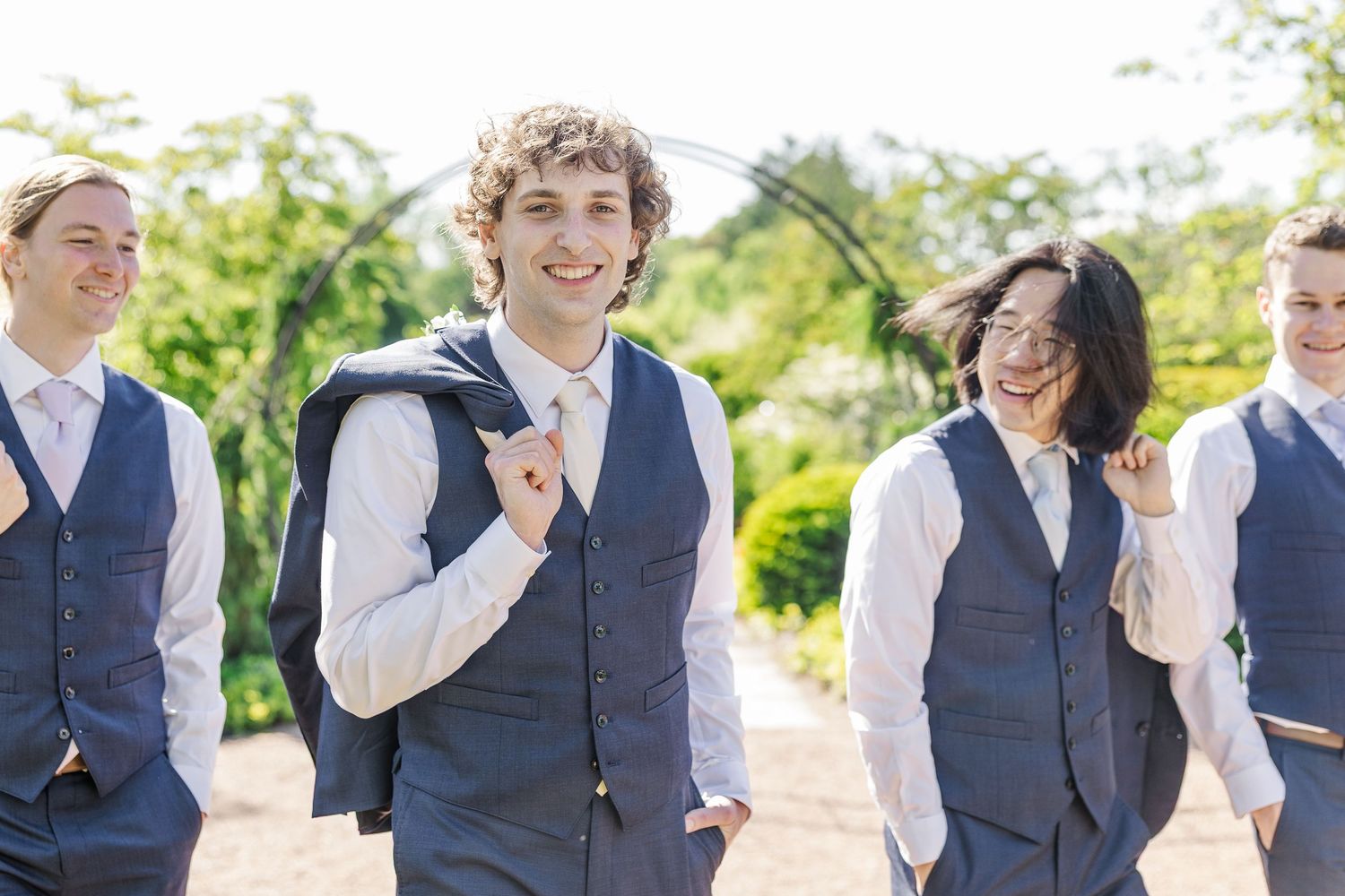 Wedding party members in matching navy blue vests walk together outdoors on a sunny day.