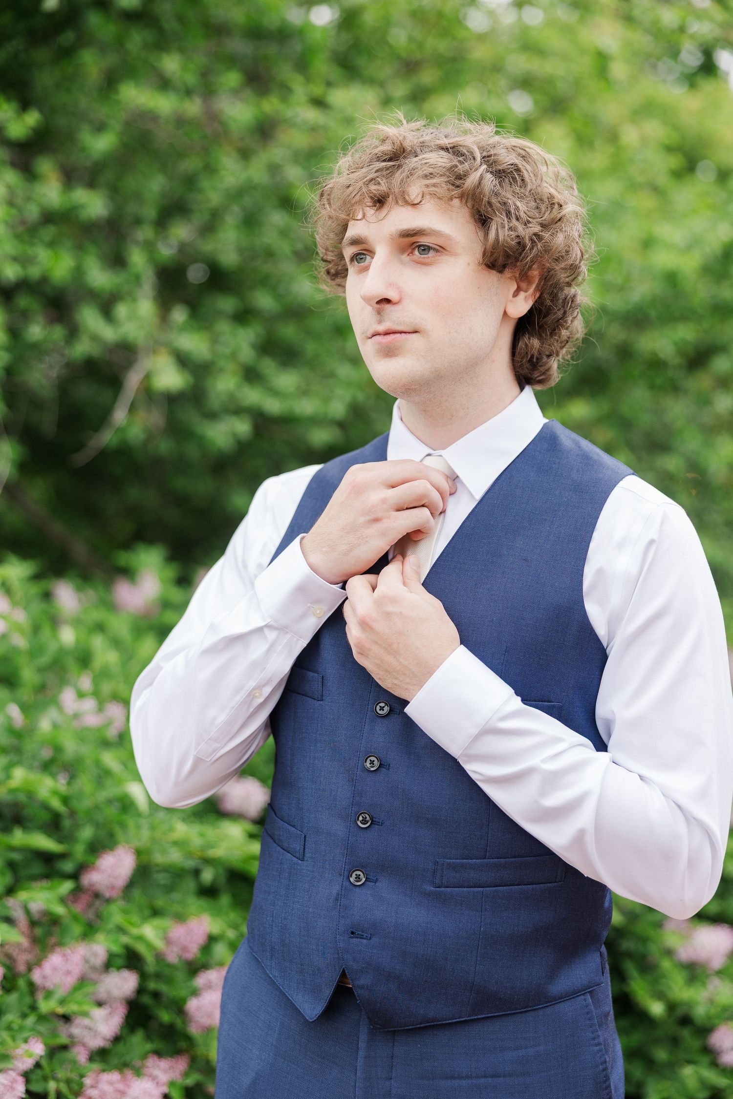 Formal sequence of someone in a navy vest and white dress shirt against a lush green garden background.