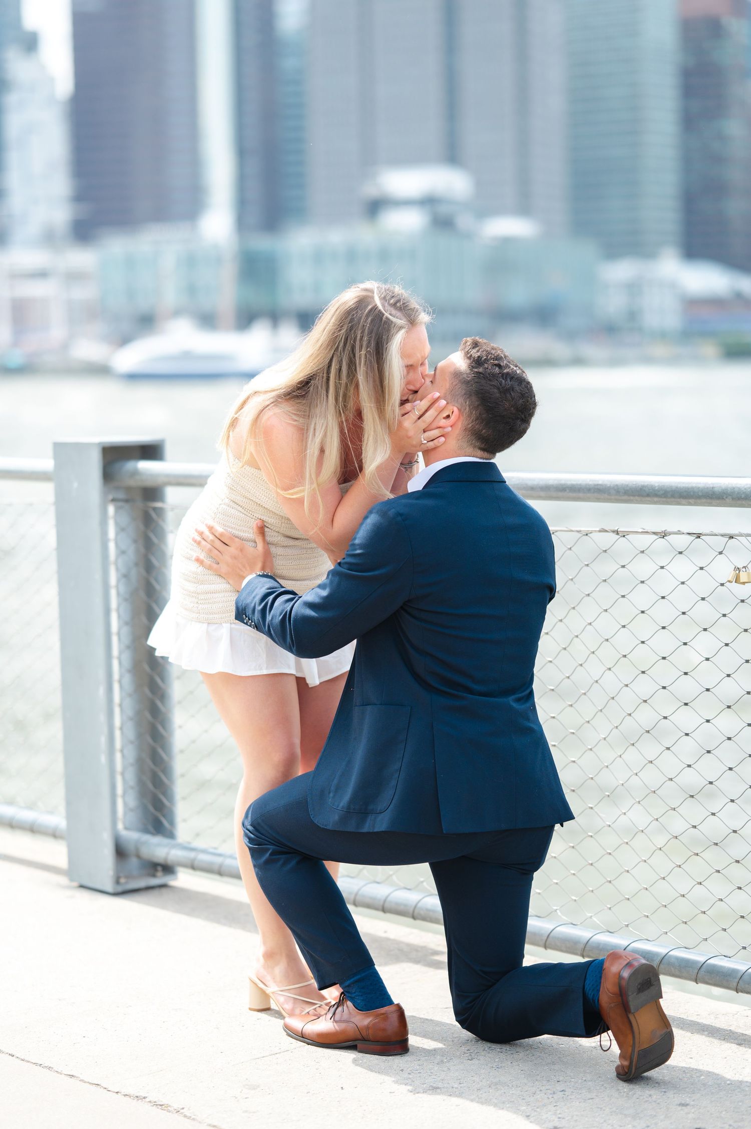 Surprise proposal moment at the Brooklyn Bridge waterfront with a cityscape backdrop as a man in a blue suit kneels down and they kiss.