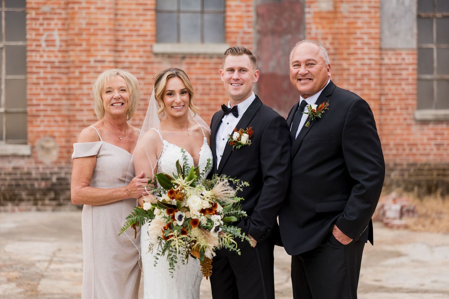 Four people in formal wedding attire standing together against a brick building with one holding a luxurious floral bouquet.