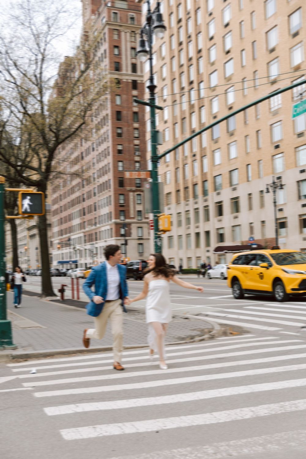 A couple walking across a crosswalk in New York City with tall apartment buildings and a yellow taxi in the background.