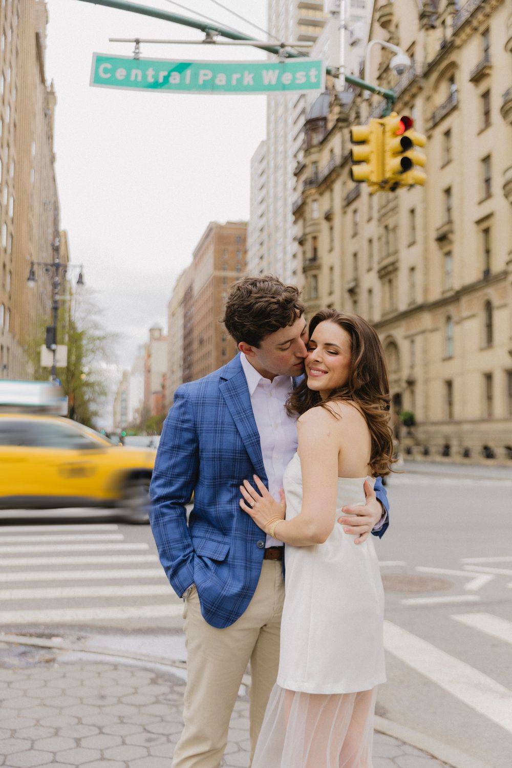 A romantic couple embraces on a Central Park West street corner in New York City with a yellow taxi passing by.