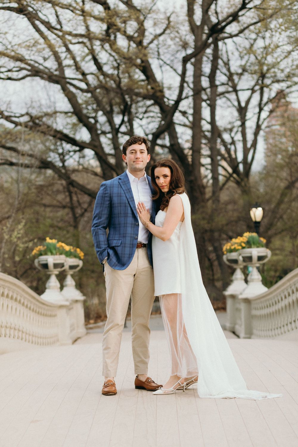 Couple in formal attire embracing on an elegant stone bridge with ornate railings and winter trees in background.