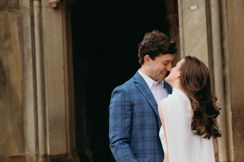 A couple shares an intimate moment outdoors, with one wearing a blue checkered suit and the other in white.
