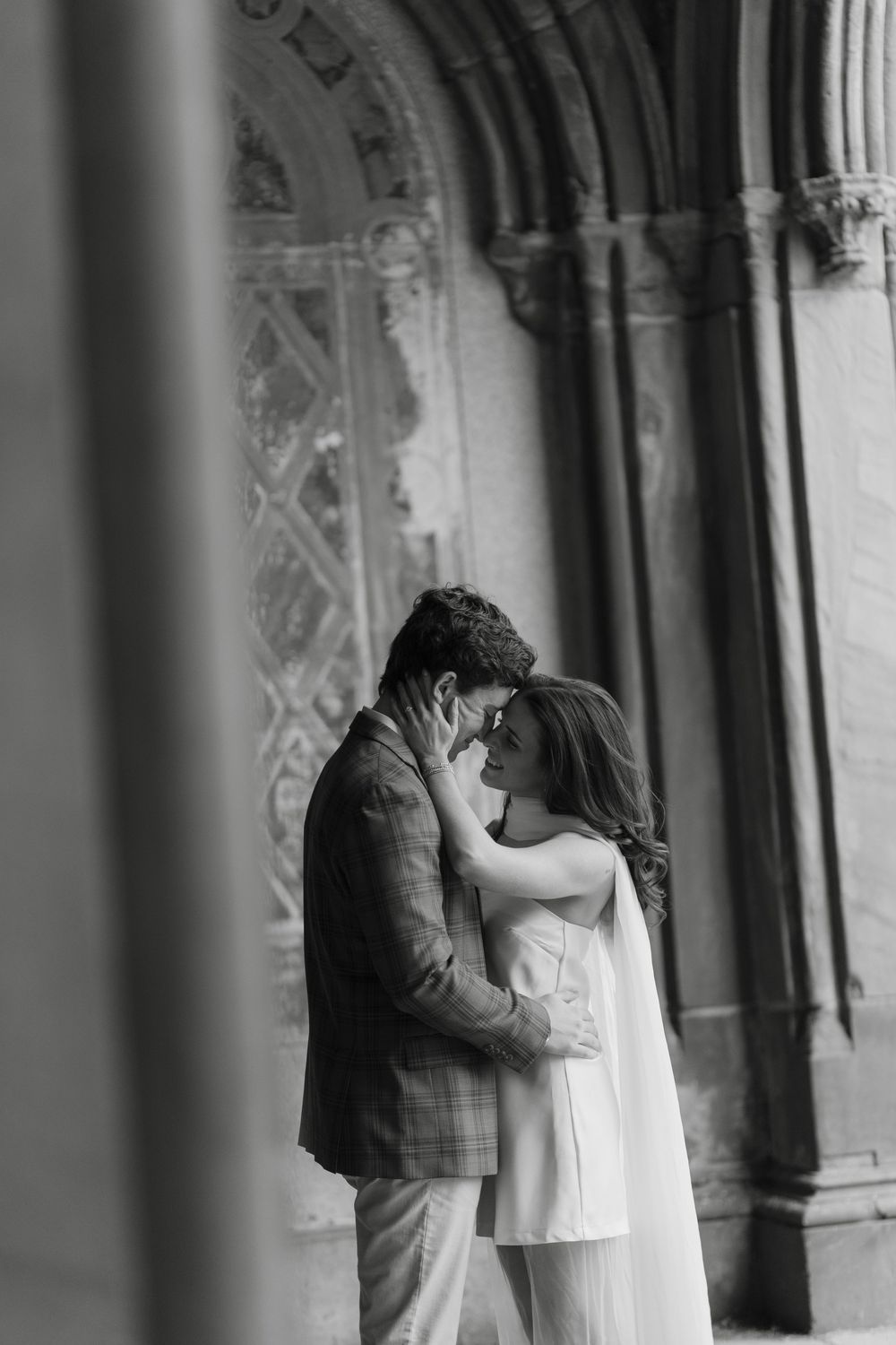 A romantic black and white photo of a couple embracing tenderly beneath ornate Gothic architecture.