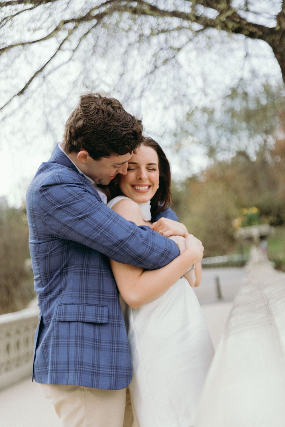 A tender embrace between a couple on a bridge, with him wearing a blue plaid blazer and her in white.