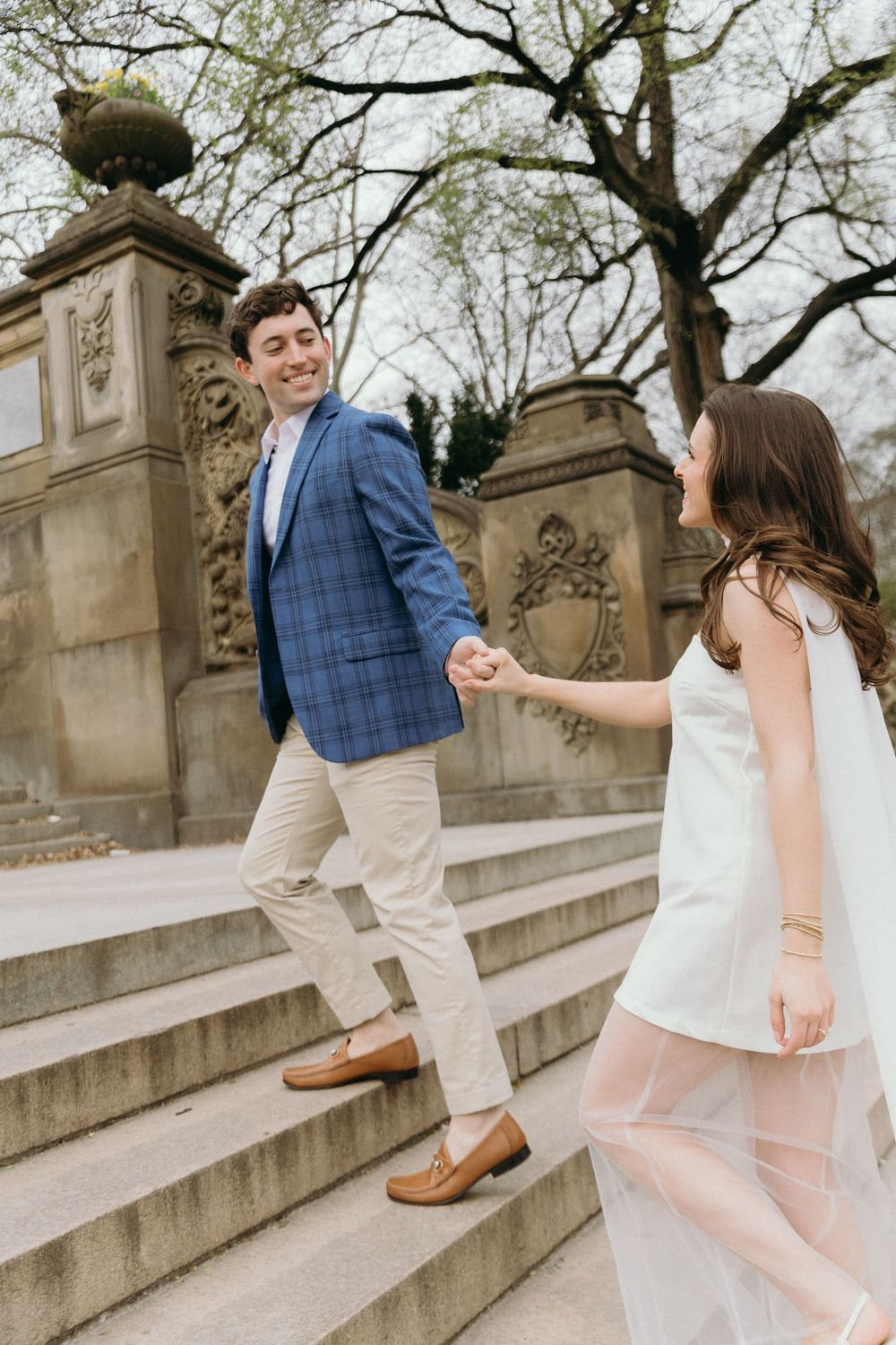 A joyful couple ascending stone steps with him wearing a blue plaid blazer and her in a flowing white dress.