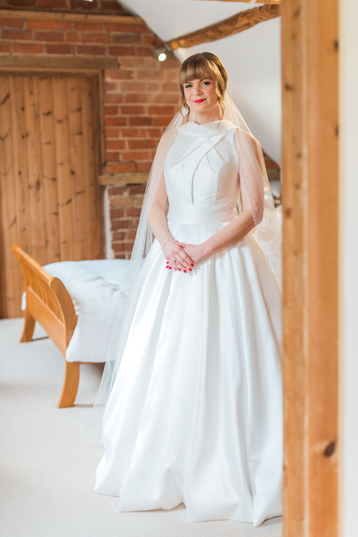 A bride in a timeless wedding dress and veil stands in a converted barn with brick walls and wooden beams ready for her cotswolds wedding