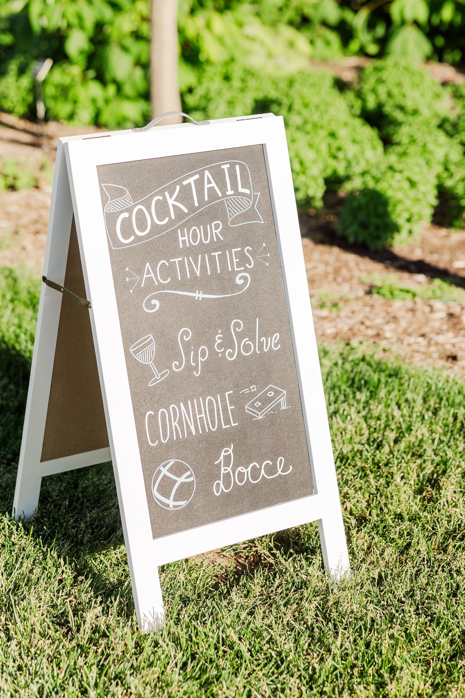 A white A-frame chalkboard sign on grass displays cocktail hour activities including sip & solve, cornhole and bocce.