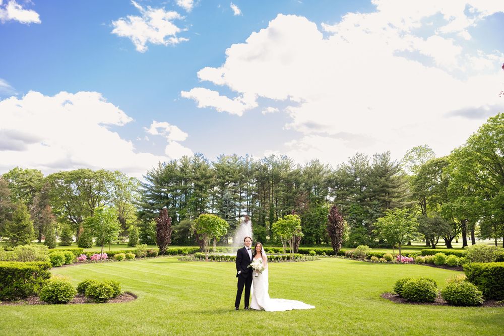 A couple stands together on a manicured lawn garden surrounded by evergreen trees on a beautiful sunny day.