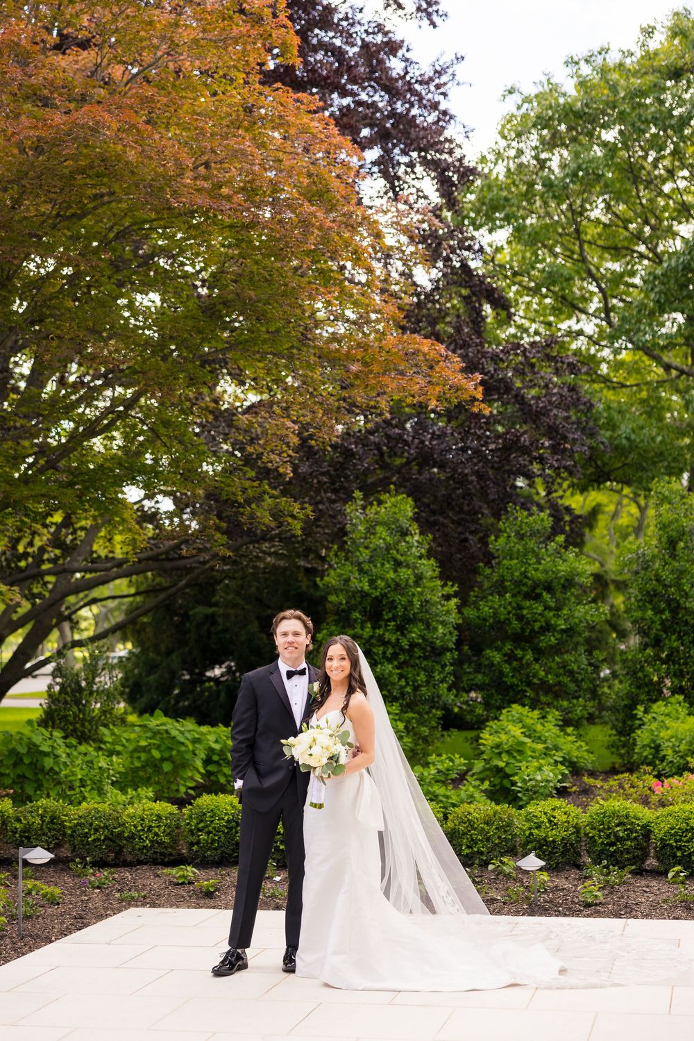A couple poses together at their outdoor garden wedding ceremony surrounded by autumn foliage and trees.