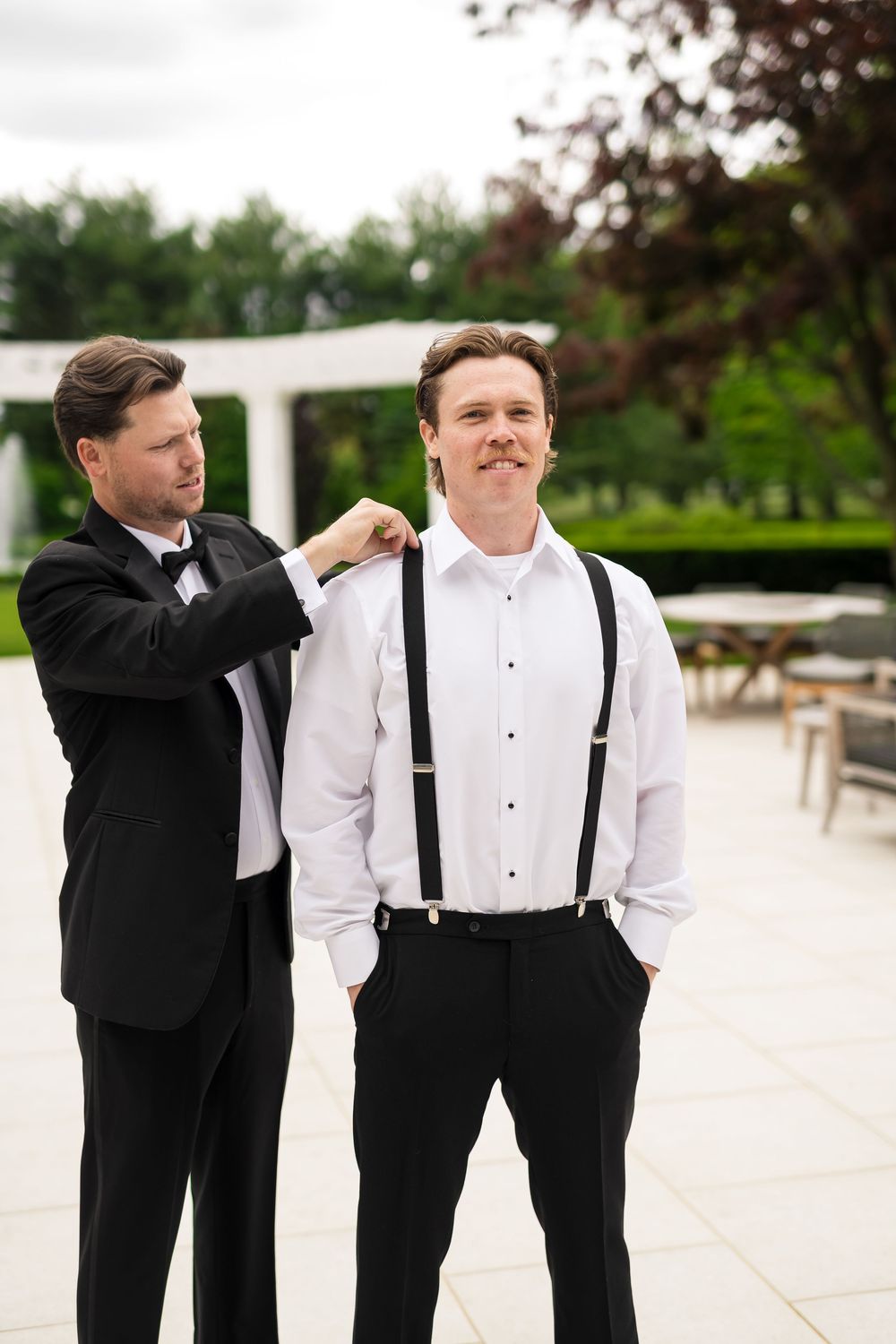 Two groomsmen in formal attire preparing for a wedding with one adjusting the other's outfit on an outdoor patio.