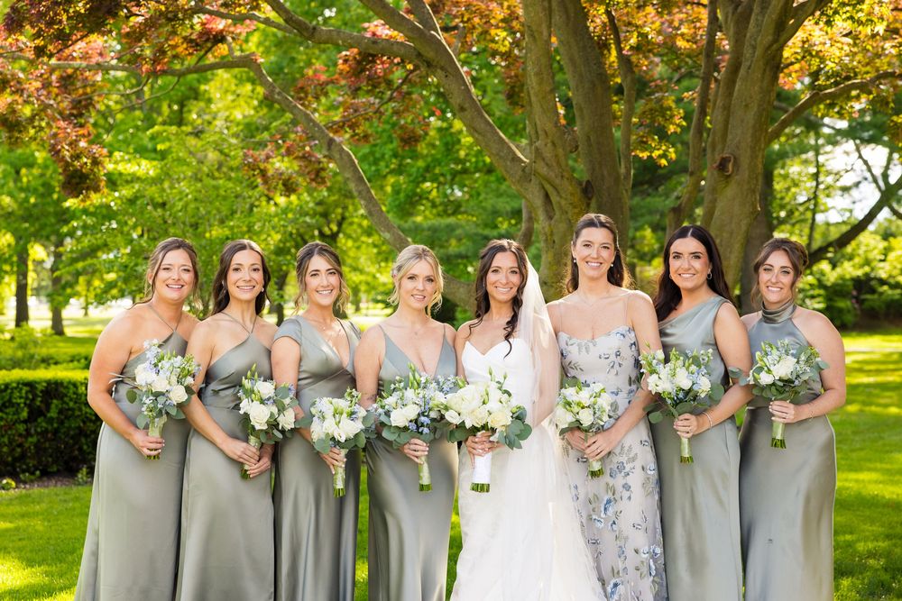 A bridal party in flowing gray dresses poses together holding bouquets in a lush garden setting.