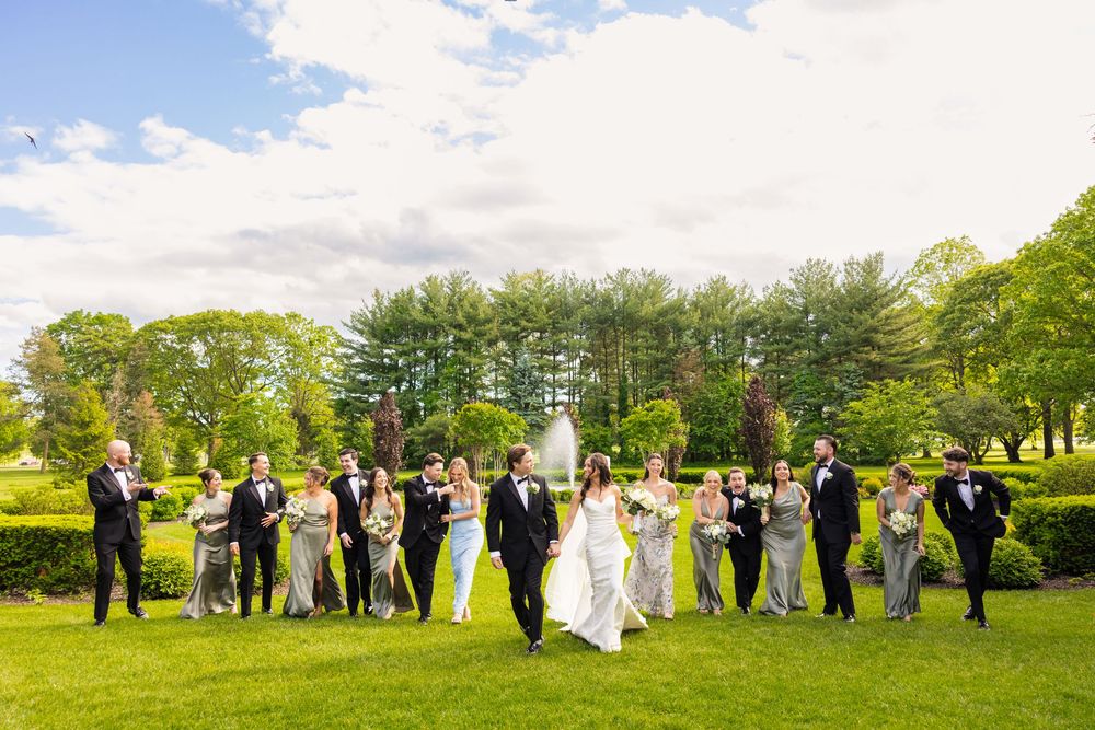 Wedding party celebrates on a lush green lawn surrounded by trees during a bright sunny day.