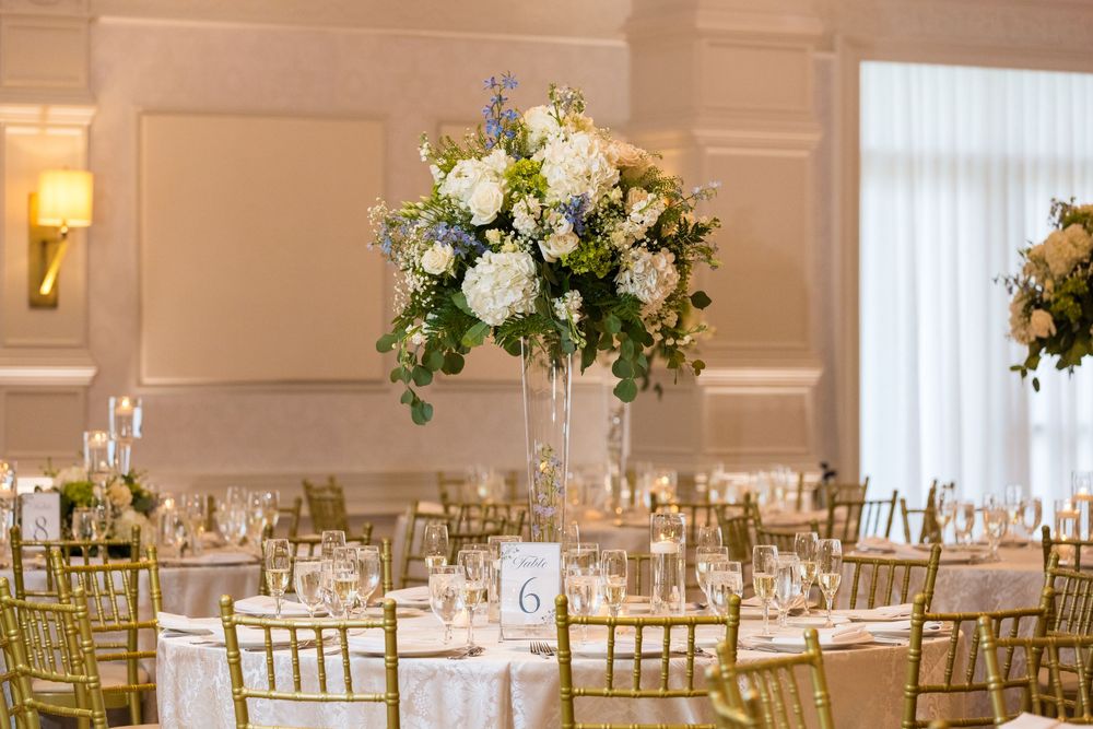 Elegant wedding reception table settings with tall white floral centerpieces and gold chiavari chairs in a ballroom.