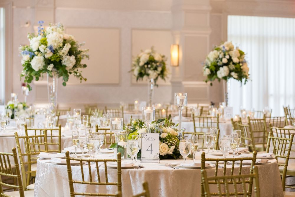 Elegant wedding reception ballroom with gold chiavari chairs and tall white floral centerpieces on round tables.