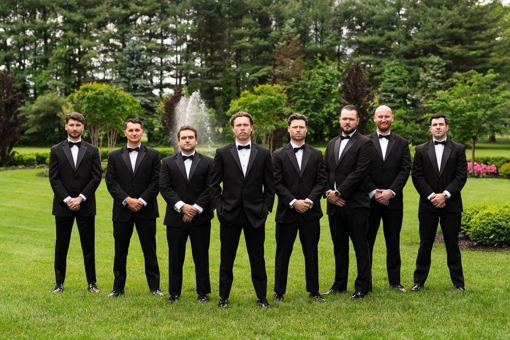 A row of groomsmen in black tuxedos pose together on a lush green lawn outside during a wedding.