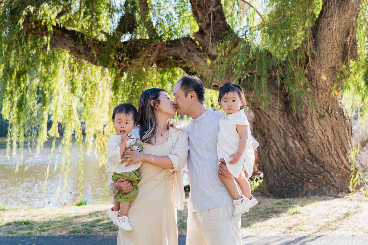 Family in white outfits poses for photo session under weeping willow tree branches in a sunlit park setting.
