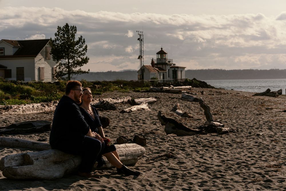 A lighthouse stands on a rocky shoreline with people sitting on the beach enjoying the coastal landscape at sunset.