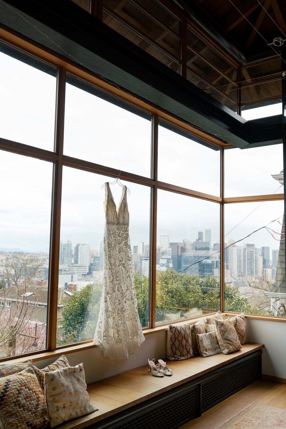 A wedding dress hangs near a window bench with throw pillows, overlooking a scenic city skyline view.