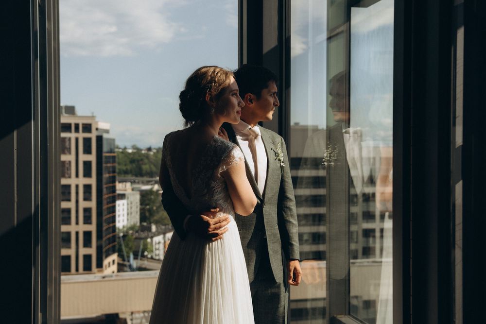 A couple in wedding attire stands together near large windows overlooking a cityscape on a sunny day.