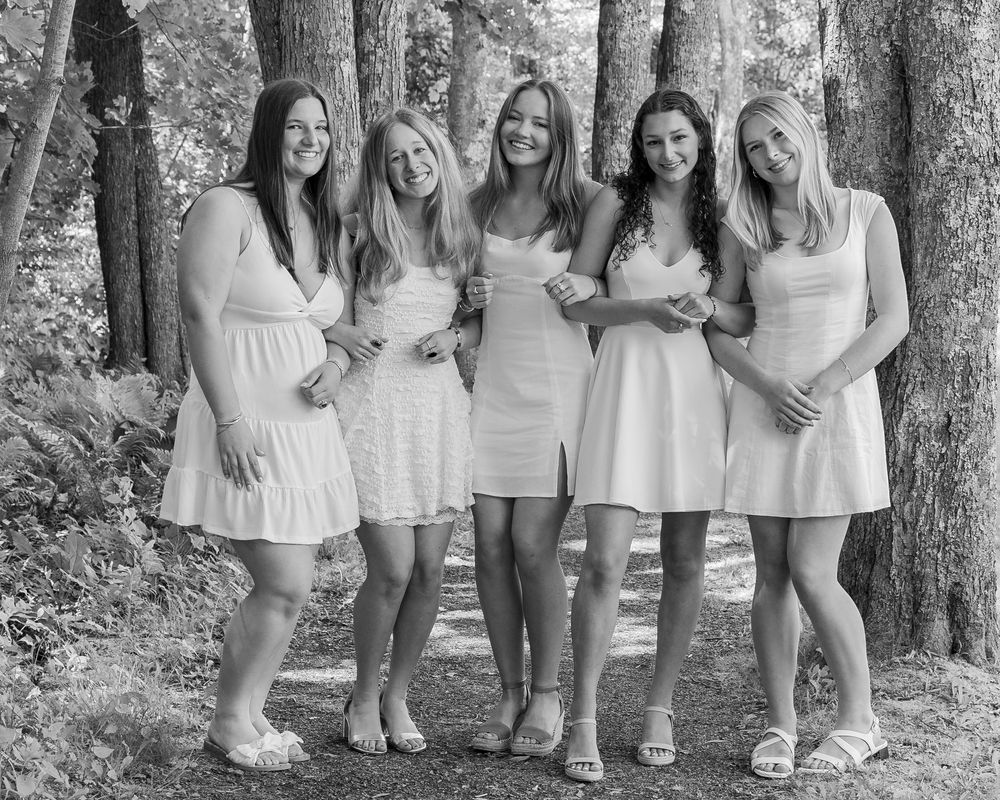 Five friends in white dresses pose together for a casual outdoor summer photo in a wooded setting.