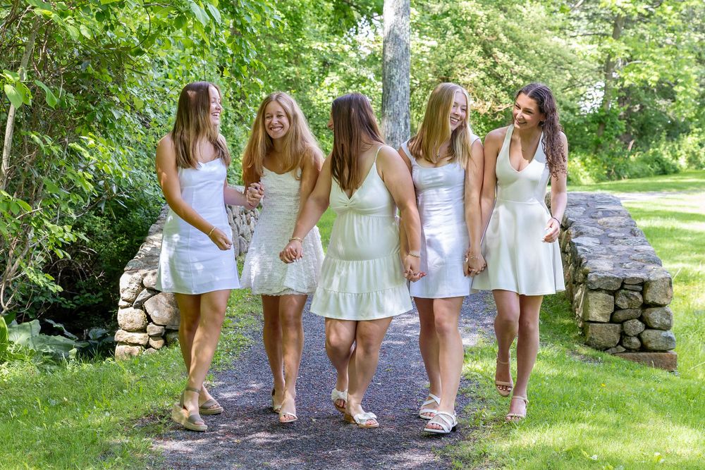 A group wearing coordinated white summer dresses walks together on a garden path bordered by greenery and stone walls.