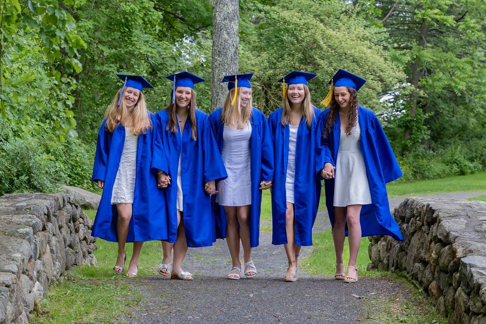 Five graduates in bright blue caps and gowns pose together on a stone bridge path surrounded by green foliage.