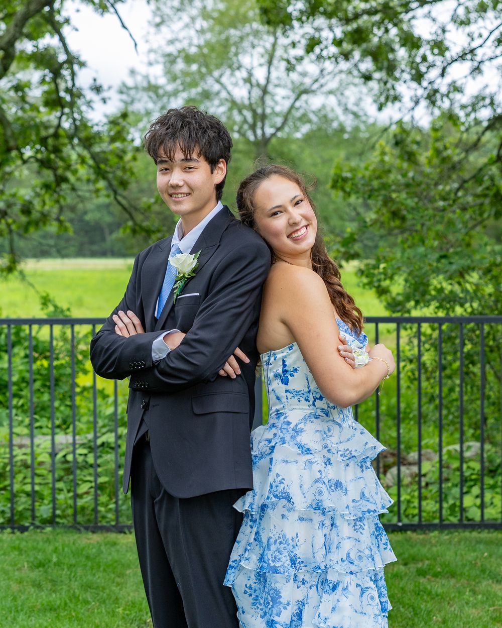 A couple poses back-to-back outdoors with a black suit and blue floral dress against green garden scenery.