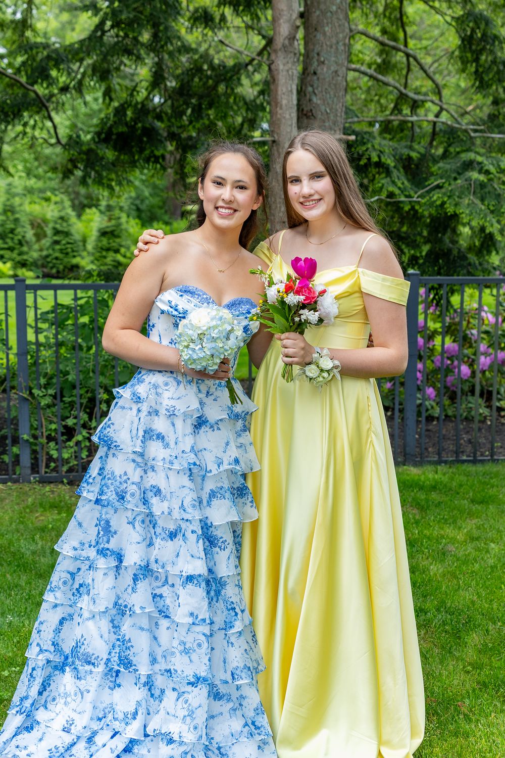Two girls in formal dresses, one in blue floral and one in yellow, pose with flowers in a garden setting.
