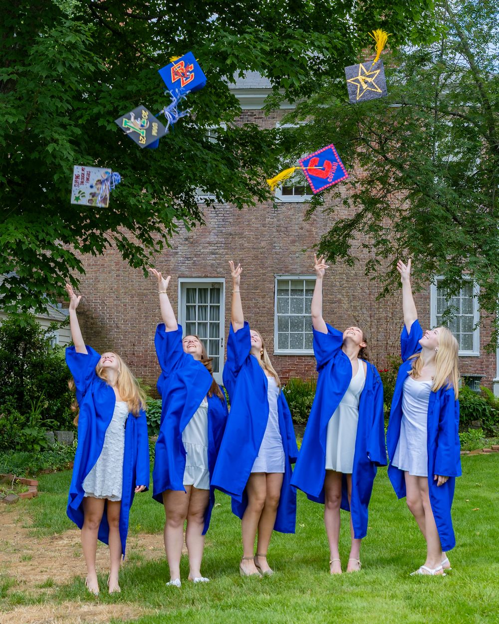 Graduates in bright blue caps and gowns toss decorated graduation caps into the air against brick building and trees.