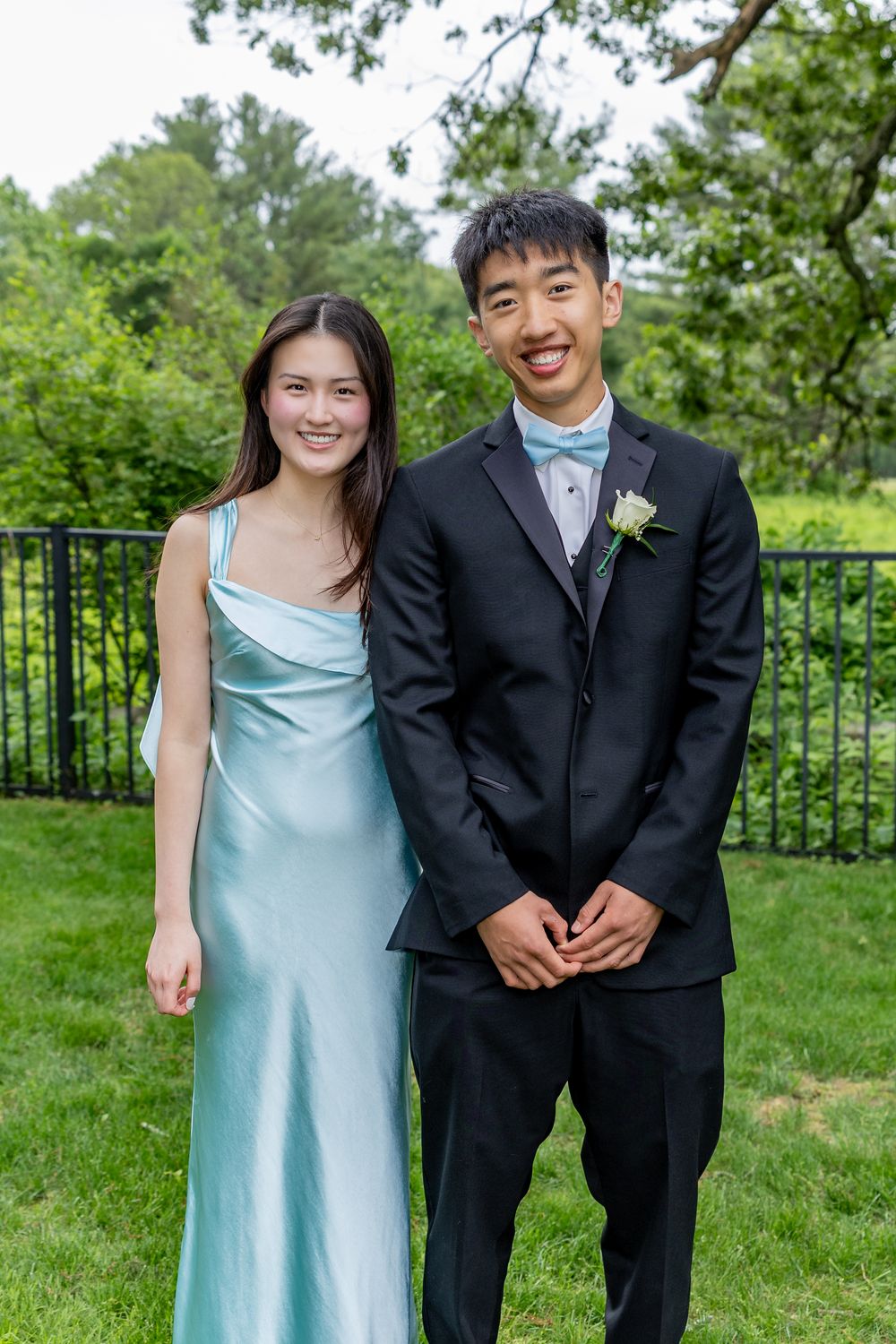 A couple in formalwear poses outdoors, with a blue satin dress and black tuxedo against a garden backdrop.